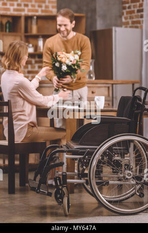 Nahaufnahme der Rollstuhl und lächelnden Mann Blumen zu präsentieren behinderte Frau zu Hause Stockfoto