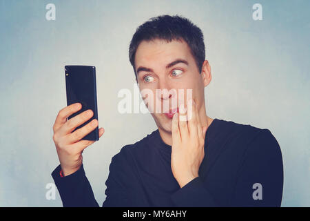 Closeup Portrait von gutaussehenden jungen Mann überrascht, schockiert, offenen Mund und Augen, durch das, was er sieht auf seinem Handy, auf blauem Hintergrund. Negative menschliche e Stockfoto