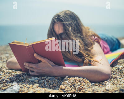 Eine junge Frau ist am Strand liegen und ein Buch lesen, an einem sonnigen Tag im Sommer Stockfoto