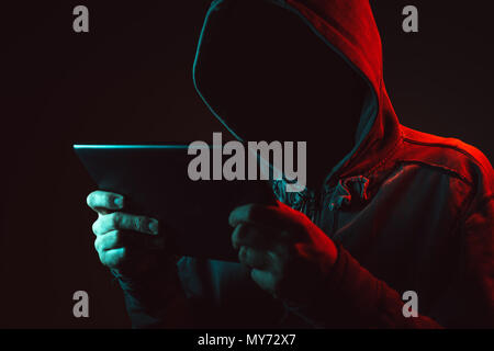 Gesichtslosen Mann mit Kapuze holding Tablet-PC im dunklen Zimmer, blauen und roten Licht low-key-Bild Stockfoto