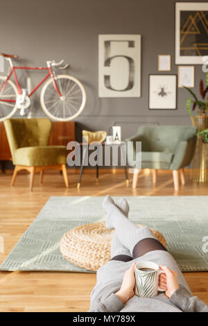 Junge Dame lag auf einem grünen Teppich mit einer Tasse Tee in der Hand und den Beinen auf einem Hocker in einem vintage Wohnzimmer Interieur mit verschwommenen Hintergrund Stockfoto