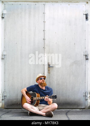 Foto eines jungen männlichen Erwachsenen das Rauchen einer Zigarre und eine akustische Gitarre spielen auf der Straße. Stockfoto