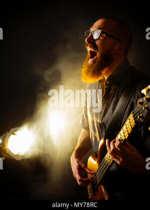 Foto von einem schreienden Bass Player mit einem Bart spielen in einem dunklen Club mit Spotlight im Hintergrund. Stockfoto