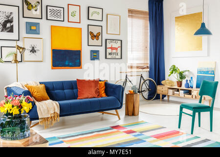 Geräumiges Wohnzimmer mit einer Decke und orange Kissen auf ein blaues Sofa, grünen Stuhl, bunten Teppich und Galerie der Plakate und Gemälde auf Weiß Stockfoto