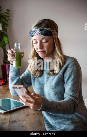 Junge Frau mit Gemüsesaft auf Smartphone im Cafe Fensterplatz