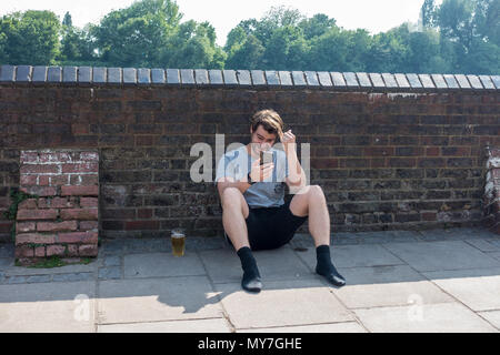 Ein junger erwachsener Mann sitzt auf dem Boden mit einem Pint Bier und konzentriert sich auf sein Mobiltelefon. Stockfoto