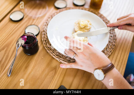 Hände einer Frau Verbreitung Creme auf einem Scone, mit Marmelade bereit, um an der Spitze zu verbreiten Stockfoto