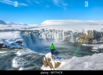 Junge Mann, der auf einem Felsen, Wasserfall mit Regenbogen, Góðafoss, Godafoss im Winter mit Schnee und Eis, Norðurland djupivogur Stockfoto