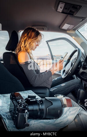 Junge Frau im Fahrzeug sitzen, bei digital Tablet suchen, SLR-Kamera auf Beifahrersitz, Mexican Hat, Utah, USA Stockfoto