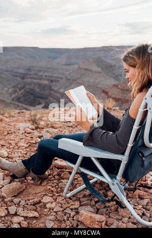 Junge Frau in remote Einstellung, auf Camping Stuhl, lesen Buch sitzen, Mexican Hat, Utah, USA Stockfoto