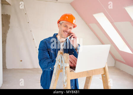Portrait von Happy Bauarbeiter mit Handy und Beratung mit jemand während mit Laptop auf der Baustelle. Stockfoto