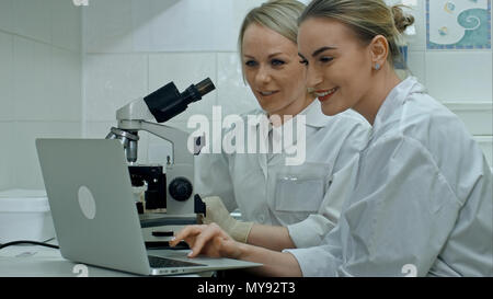 Zwei junge positive Wissenschaftler im Labor arbeiten mit Mikroskop und Laptop Stockfoto