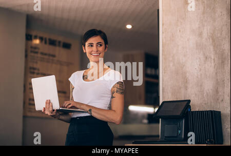 Lächelnd kaukasischen stehende Frau in ihrem Coffee Shop mit einem Laptop. Weibliche Manager mit Laptop in ein Restaurant. Stockfoto