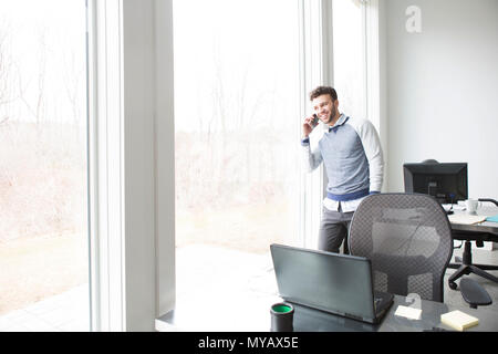 Ein junger Geschäftsmann schaut aus einem Büro Fenster beim Chatten auf ein Handy. Stockfoto