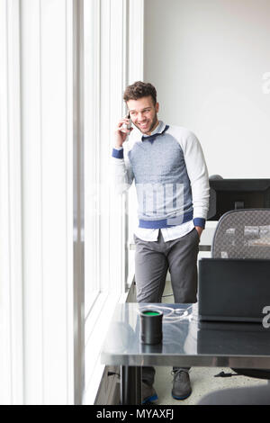 Ein junger Geschäftsmann schaut aus einem Büro Fenster beim Chatten auf ein Handy. Stockfoto