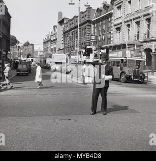 1960, historische, tagsüber und ein uniformierter Britischer Polizist mit Hemet gesehen Leitet den Verkehr in der Mitte einer Straße in London, England, UK. Stockfoto