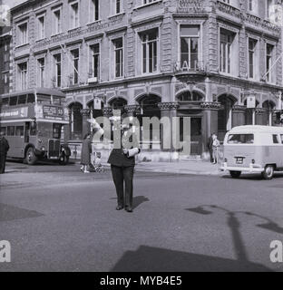 1960, historisch, tagsüber und ein uniformierter britischer Polizist mit Helm wird gesehen, wie er den Verkehr in der Mitte einer Londoner Straße, England, führt. Stockfoto