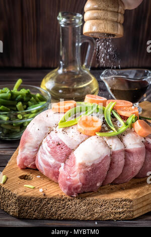 Rohes Schweinefleisch chop Fleisch Rollen aufgewickelte Garn Jute marinierte Streuen grobes Meersalz Shaker Mill Olivenöl mit Pfeffer verschiedene Gemüse mit Möhren grün auf Stockfoto