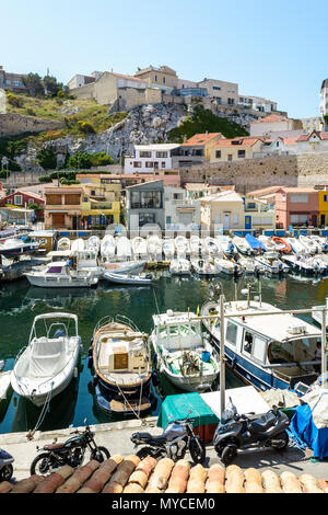 Der kleine Hafen von Endoume in Marseille durch einen sonnigen Morgen mit Booten vor bunten cabanons und Häuser unterhalb der Klippe vertäut. Stockfoto