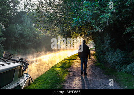 Trent & Mersey Canal, Derbyshire. UK Wetter. 07.06.2018. Nebel, dunstiger Atmosphäre Start in den Tag Für ein Hausboot Bewohner nehmen am Morgen schlendern. Die stimmungsvolle Szene durch den Dampf hat steigende die Oberfläche dieser Sonne beleuchteten Kanal ist eine, die häufiger bei schlechtem Wetter verschiebt sich vom warmen Sommer Sonnenaufgänge zu klaren, kalten Morgen. Das Phänomen hat viele Namen, einschließlich Dampf Nebel, Verdunstung Nebel, Frost-, Rauch- und Meer rauchen. Stockfoto