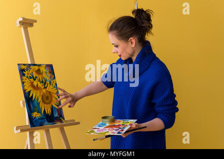 Mädchen Künstler malt Sonnenblumen Ölfarben auf Leinwand. Sie trägt eine blaue Pullover. Frau hält Pinsel und Palette mit Farben. Sie setzt Farbe Stockfoto