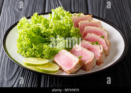 Leckere scheiben Thunfisch Steak gebraten in Panade mit Blattsalat und Kalk closeup auf einer Platte auf einem Schwarzen Tisch serviert. Horizontale Stockfoto