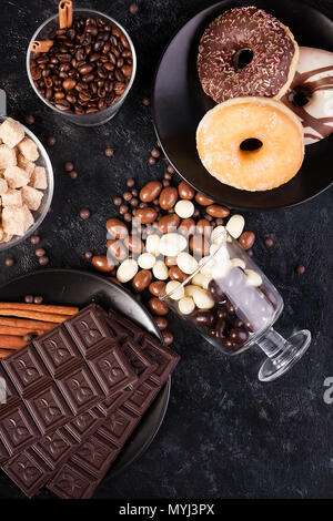 Blick von oben auf die Erdnüsse in Schokolade, auf der dunklen Kammer verschüttet, neben Schokolade Tabletten, Donuts, brauner Zucker und Kaffee Bohnen Stockfoto