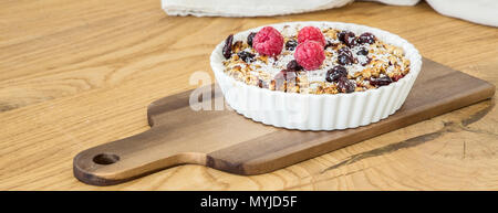 Tiefe weiße Platte mit Müsli, getrocknete Cranberries, Nüsse, frisch Himbeeren und Kokosnuss auf einem Brett auf hölzernen Tisch Stockfoto