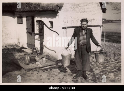 Ein dänischer Bauer Landwirt, circa 1925, außerhalb seiner Reetdachhaus. Er trägt zwei Eimer von einem Joch auf seinem Rücken, wahrscheinlich mit Wasser aus der alten Pumpe direkt hinter ihm; er trägt Jacke, T-Shirt und ein Rohr, wie er lächelt für sein Portrait genommen zu werden. Stockfoto