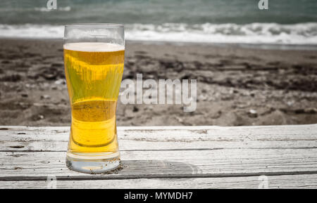 Glas Licht golden Pilsner Bier auf einem rustikalen Tisch draußen auf dem Sand am Meer mit dunklem Sand und das Brechen von Brandung im Hintergrund Stockfoto