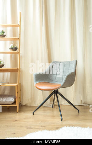 Grau Designer Stuhl gegen einen Vorhang, neben Holzregals im Wohnzimmer mit weißem Pelz auf dem Boden Stockfoto