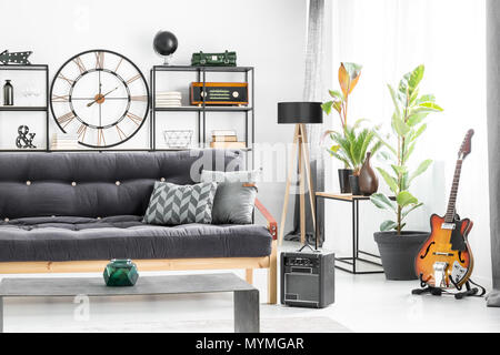 Grau Kissen auf dunklen Sofa vor eine Tabelle in der man Wohnzimmer Interieur mit Gitarre und Designer Uhr an der Wand Stockfoto