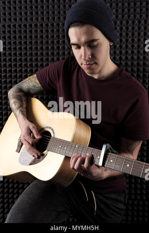Ein Kerl mit Tätowierungen in einem dunklen T-Shirt und Hut spielt auf der Gitarre. Vertikale Rahmen Stockfoto
