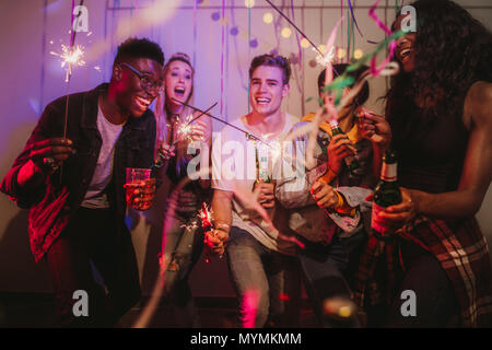 Freunde feiern mit Feuer glitzert und Getränke an einer Party. Junge Männer und Frauen Spaß bei einem bunten House Party mit Dekorationen und conf in Stockfoto