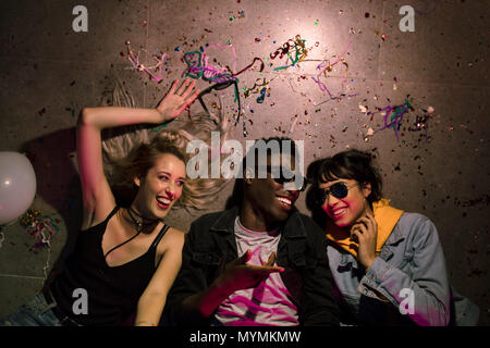 Blick von oben auf die Mann und zwei Frauen liegen auf dem Boden und Lachen an einem Haus Party. Freunde liegen auf dem Boden an der House Party mit Ballons und Konfetti um Stockfoto
