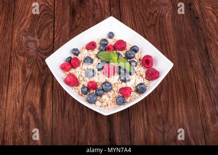 Weiße Schüssel voll mit Hafermehl mit Himbeeren, Heidelbeeren und Minze auf einem Holztisch Stockfoto