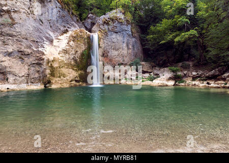 Ein friedlicher Wasserfall, der in einen kristallklaren, smaragdgrünen Pool stürzt, umgeben von felsigen Klippen und üppigem Grün, Stockfoto