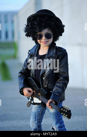 Eine kleine, niedliche junge Gitarristen in Lederjacke und Sonnenbrille, Gitarre zu spielen. Junge Idol. Kinder- Interessen und Hobbys. Sein wie ein Rockstar. Stockfoto