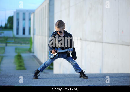 Eine kleine, niedliche junge Gitarristen in Lederjacke und Sonnenbrille, Gitarre zu spielen. Junge Idol. Kinder- Interessen und Hobbys. Sein wie ein Rockstar. Stockfoto