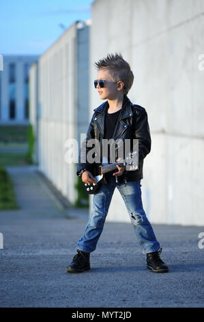 Eine kleine, niedliche junge Gitarristen in Lederjacke und Sonnenbrille, Gitarre zu spielen. Junge Idol. Kinder- Interessen und Hobbys. Sein wie ein Rockstar. Stockfoto