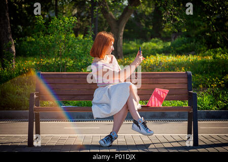 Eine junge rothaarige Frau in einem weißen romantische Rock, pink und blau Sneakers Sneakers, macht selfie am Telefon und sitzt auf einer Bank an einem Sommertag Stockfoto