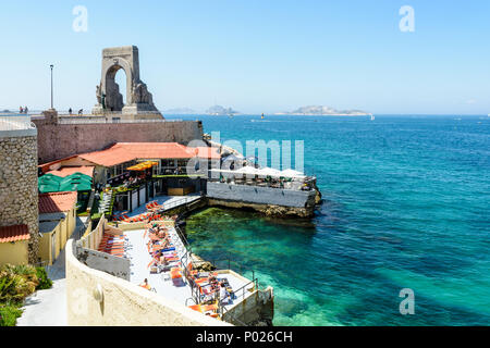 Ein Beach Restaurant am Ufer unter dem kriegerdenkmal an der Östlichen Armee in Marseille, Frankreich, hat seine Terrasse mit Sonnenschirmen und Liegestühlen. Stockfoto