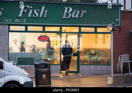 Britischer Fish & Chips Shop, Geschäftsreisenden geöffnet. Der Kunde öffnet die Tür, um sein Essen zum Mitnehmen abzuholen, ein anderer Gast sitzt drinnen und isst auf dem Gelände. Stockfoto