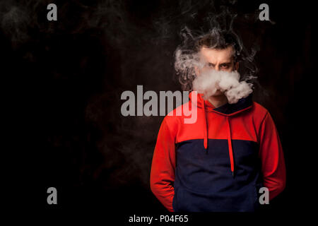 Eine junge dunkelhaarige Mann mit den traurigen Augen in einer farbigen orange und blau Hoody stehend mit Arme hinter raucht eine vape und atmet ein weißer Rauch auf einem schwarzen Stockfoto