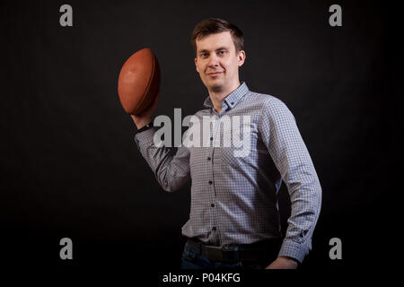 Dunkelhaarige Mann in einem Plaid Shirt hält einen Rugby Ball in seinen Händen auf schwarzem Hintergrund isoliert Stockfoto