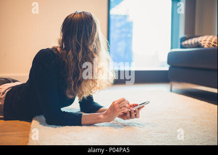 Eine junge Frau ist auf dem Boden liegend in ihrer Wohnung und ist mit einem Smartphone Stockfoto