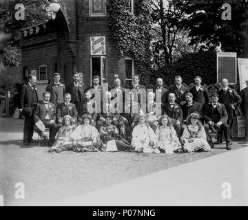 . Englisch: Eine Gruppe Portrait an Rosherville Gärten, Northfleet ein 12 x 10 negativ. Ein formell gestellt Gruppe von Frauen und Mädchen am Haupteingang des Rosherville Gärten, Northfleet. Die Männer sind alle Anzüge gekleidet und sie sind alle mit einer Rosette auf der linken Revers. Die sechs junge Damen auf dem Boden sitzend Tragen ausgefallenen und dekorativen Kostümen und halten gedruckte Programme für Veranstaltungen in den Gärten gehalten zu werden. Der Eintrittspreis ist gezeigt zu sixpence und einen Zeitplan für das South Eastern & Chatham Eisenbahn ist auf einer Wand in der Nähe eine "Lösung" zu unterzeichnen. Die Gärten waren Lai Stockfoto