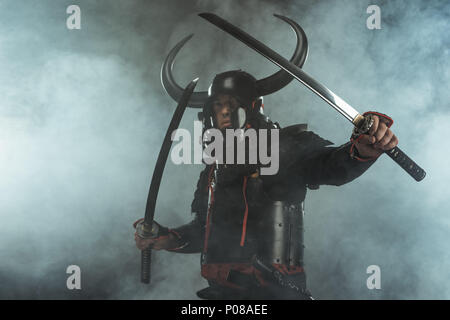 Samurai in traditioneller Rüstung mit zwei Katana Schwerter in Verteidigungsposition auf dunklem Hintergrund mit Rauch Stockfoto