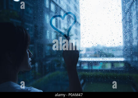 Traurig einsam liebe Frau in Regen Herz auf Fenster Glas dunkelblau Ton Stockfoto