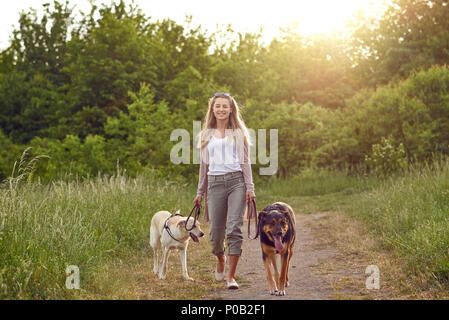 Glückliche junge Frau zu Fuß ihre Hunde entlang einer Grasbewachsenen ländlichen Track im Frühjahr mit Blick auf die Kamera Stockfoto